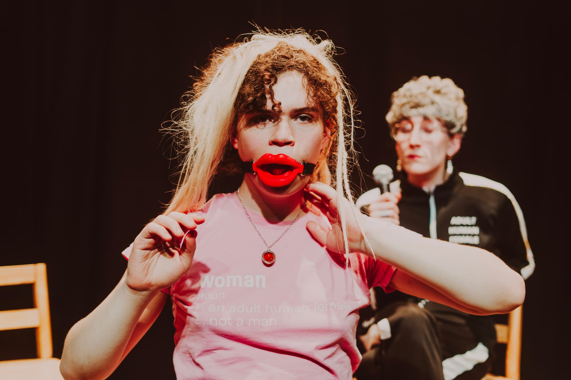 A performer with curly hair with rough blonde extensions wearing a pink t-shirt that reads 'Woman' looks out at us and wears a red mouth gag lip piece. Another performer, holding a microphone, with glasses and a black sports suit, is visible in the background. The lighting creates a warm, intimate atmosphere typical of experimental theater.