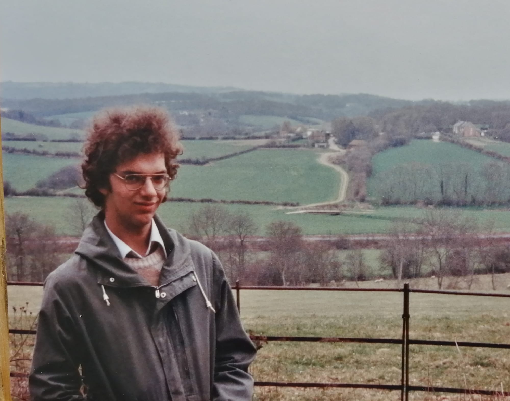 Young man with curly reddish-brown hair and glasses stands in front of a metal gate in the English countryside, circa 1980s. He wears a grey jacket over a collared shirt. Behind him stretches a pastoral landscape of rolling green fields divided by hedgerows and bare winter trees, with scattered farm buildings visible in the distance. 