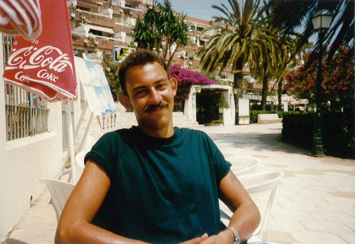 Handsome young man with short dark hair and mustache wearing teal t-shirt sits on white bench in sunny Mediterranean setting with palm trees, Coca-Cola umbrella, and apartment buildings in background.