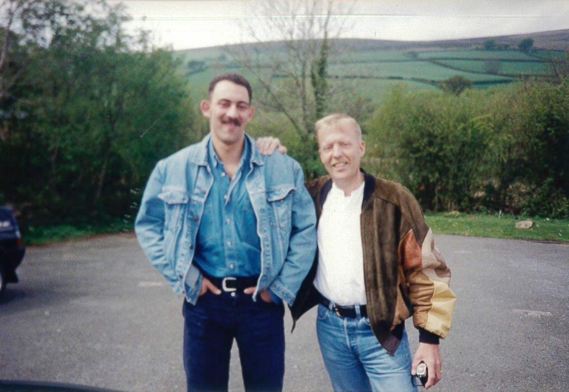 Two happy smiling gay men stand together in countryside setting, circa 1990s. Left man wears denim jacket with mustache; right man in brown bomber jacket and white shirt. Rolling green hills visible in background.