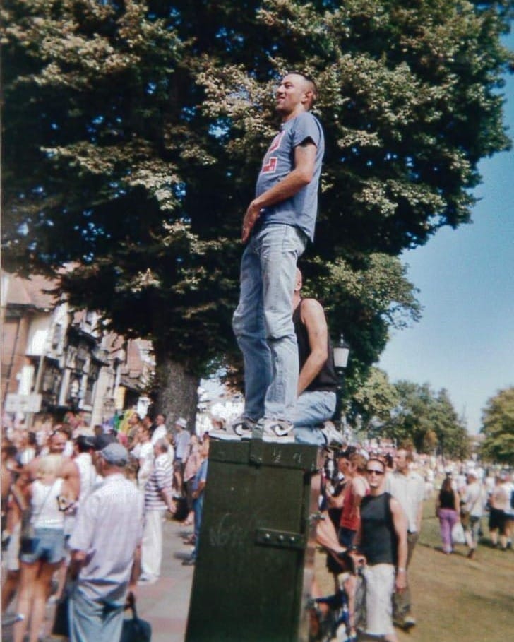 Young gay man, in jeans and t-shirt standing on a utility box in a crowded outdoor Pride event, surrounded by trees and people under a clear blue sky.