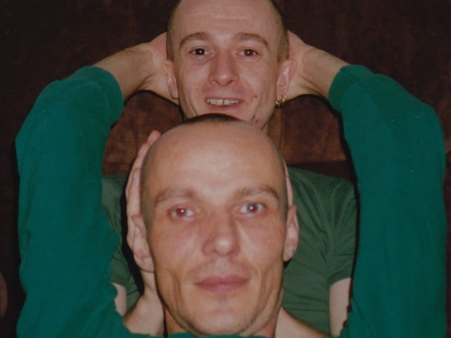 A handsome gay couple relaxing together on a brown couch, one wearing a green shirt reclining in front while the other smiles behind him, capturing an intimate moment from the 1990s.