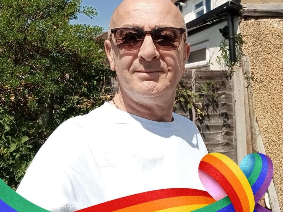 Man wearing sunglasses and white shirt outdoors with Pride rainbow flag overlay, standing in front of residential home on sunny day.