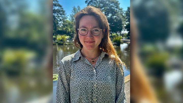 A portrait of Marina in front of a pond and greenery, smiling slightly at the camera on a sunny day