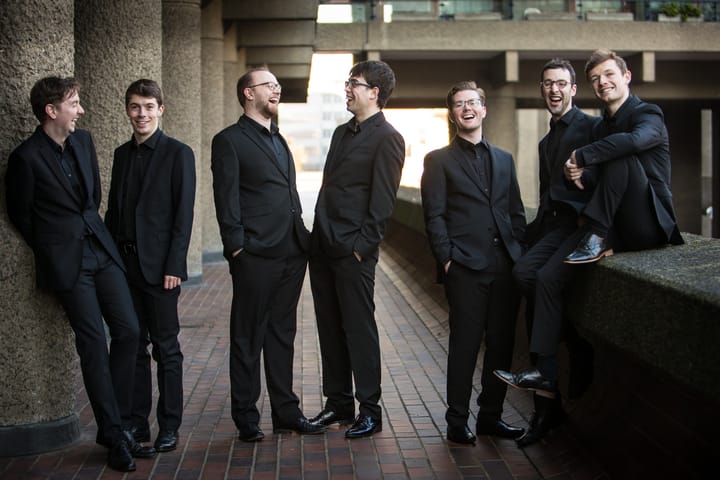 Six handsome men in black suits gathered in a brutalist concrete corridor with brick flooring and concrete pillars, engaged in conversation and smiling.