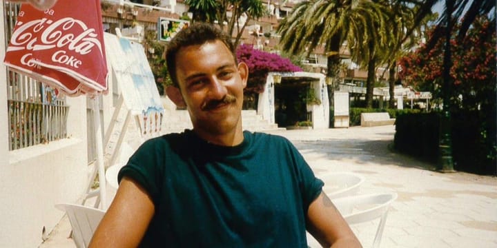 Handsome smiling young man, seated outdoors at a café under a Coca-Cola umbrella, with palm trees, vibrant bougainvillea, and terraced buildings in the sunny background.